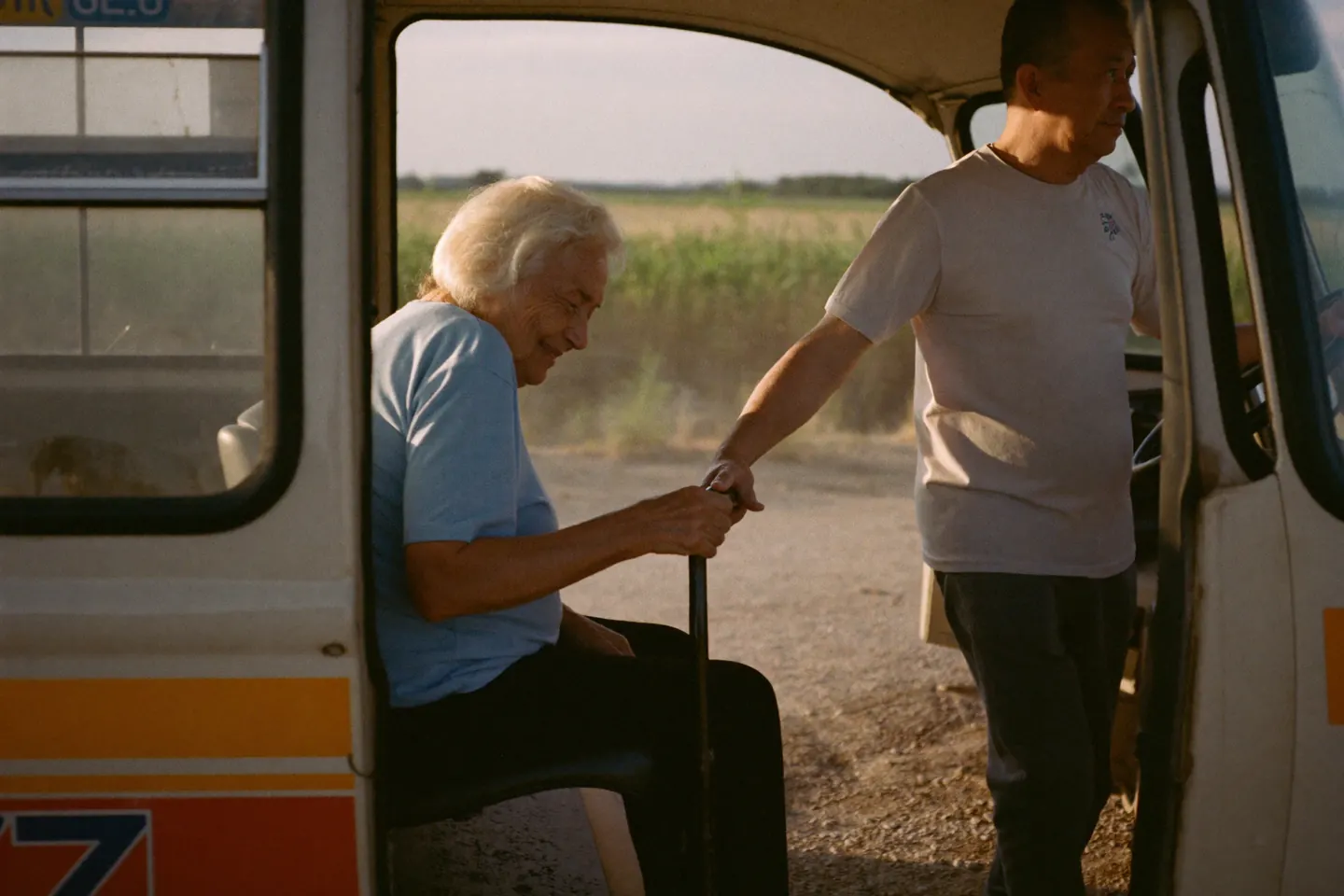 A driver steadies an elderly woman with a cane as she boards a small rural minibus at a roadside stop, late afternoon light and fields behind them.