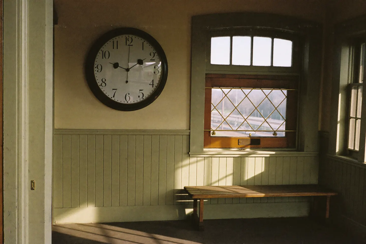 The interior of a small historic depot: a large round wall clock above wainscoting, an empty bench, and long afternoon window light across the floorboards.