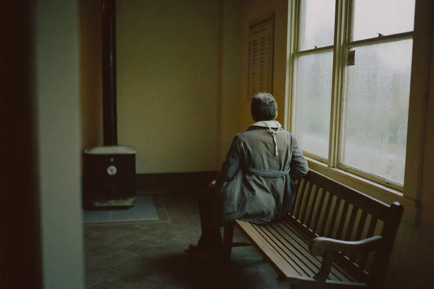 A lone passenger in a canvas coat sits on a wooden bench in an empty country depot waiting room, looking out through rain-streaked windows.