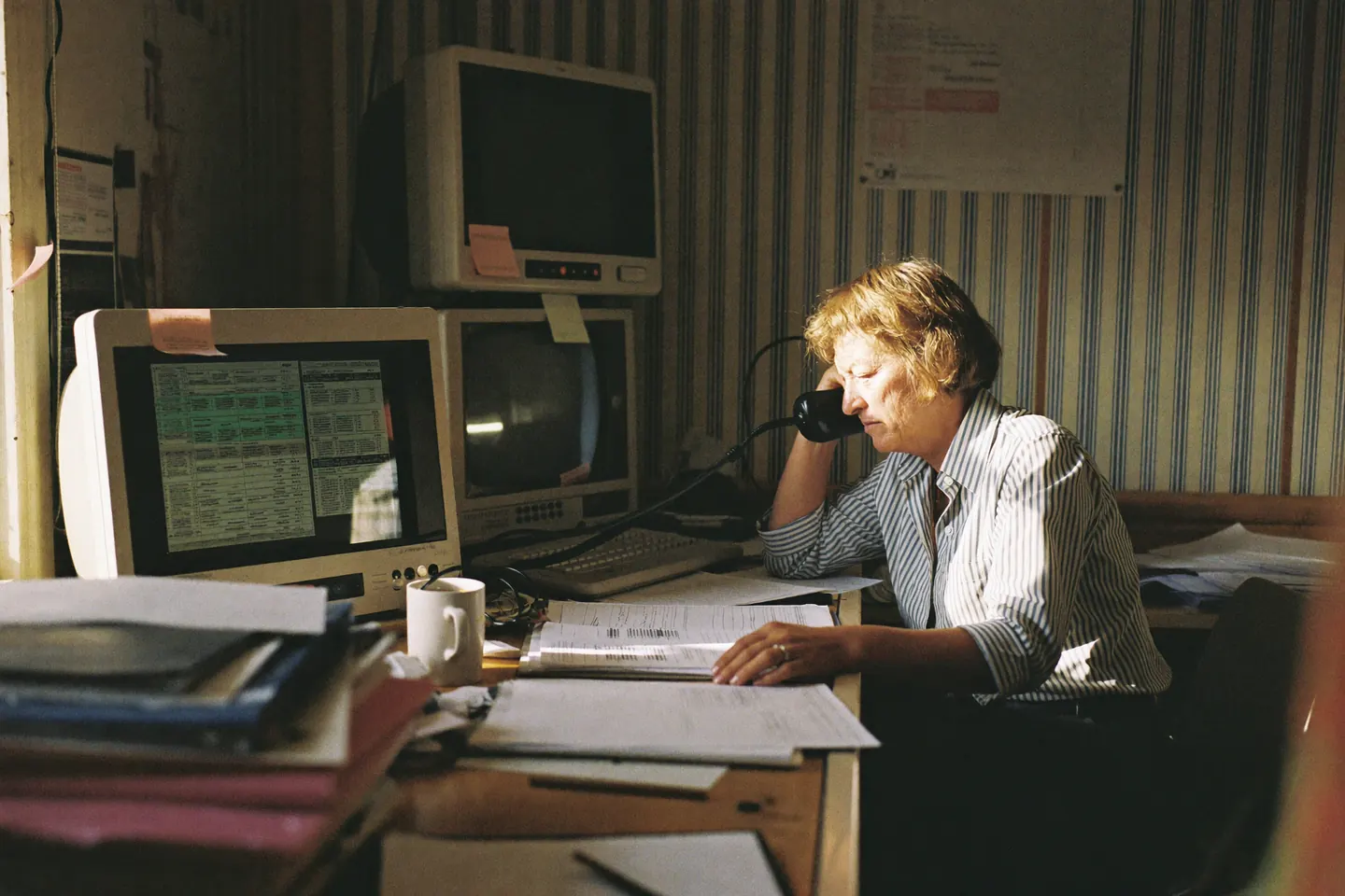 A dispatcher on the telephone at a crowded office desk, surrounded by stacked paper schedules, an older monitor and a coffee mug, lit by low morning sun.