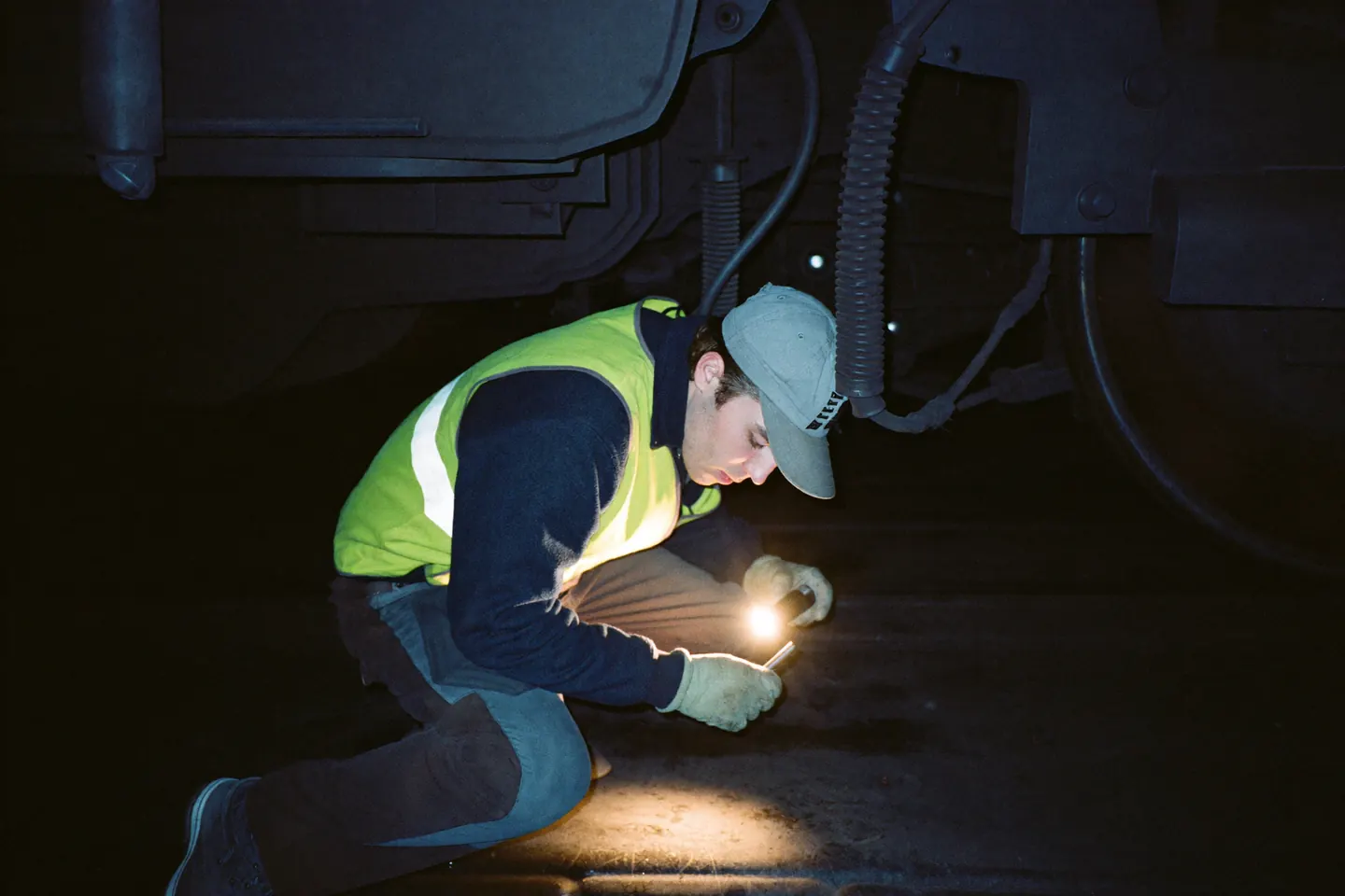 A mechanic in a hi-vis vest crouched beneath a railcar with a drop light, inspecting a brake assembly in the dark of an inspection pit.