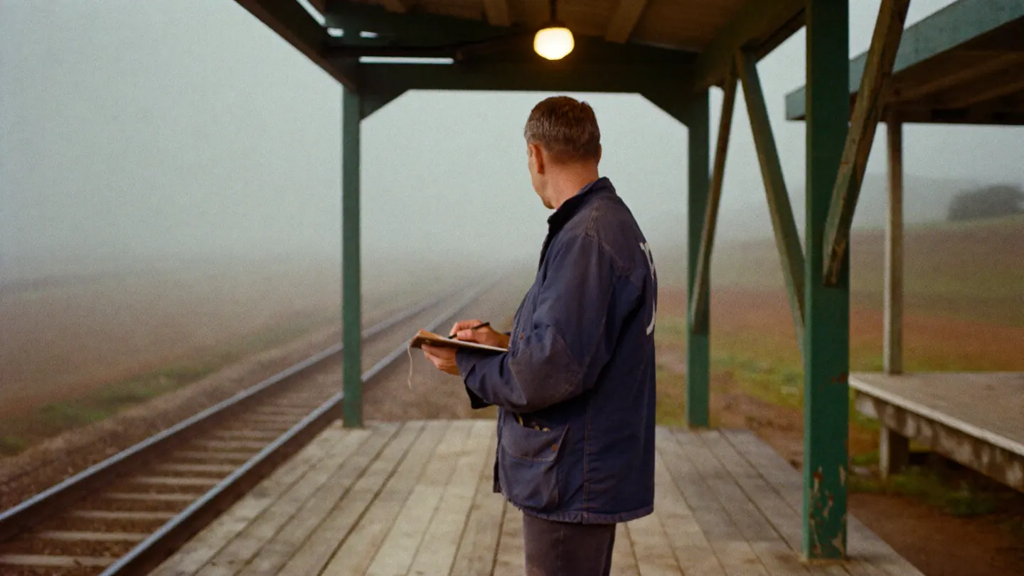 A conductor in a worn navy work jacket stands alone on a wooden rural platform in heavy morning fog, writing on a clipboard, looking down an empty single track.