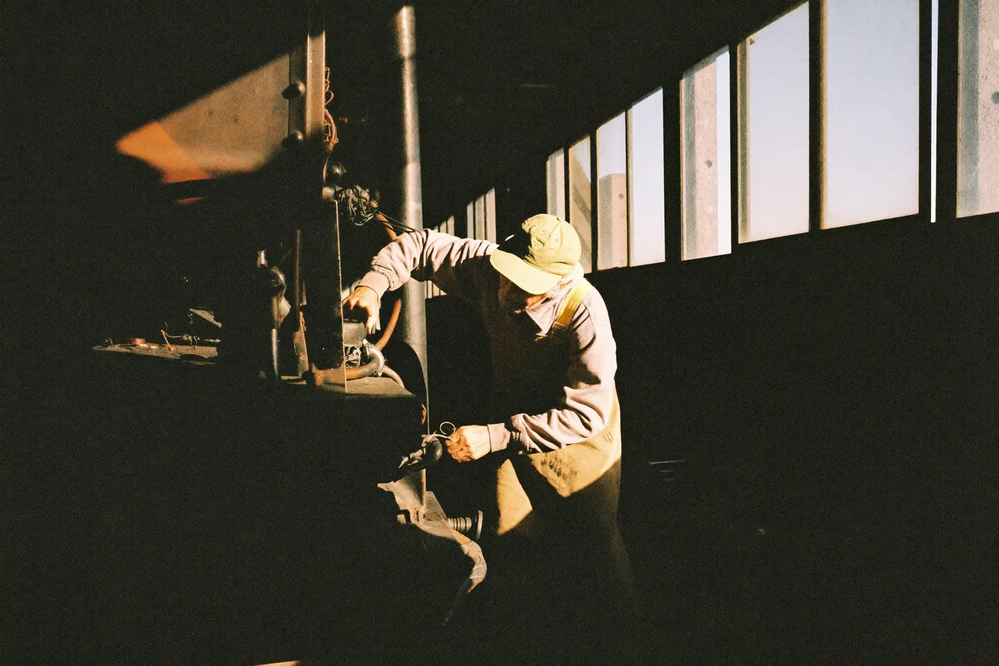 A worker in a hard hat silhouetted against tall shed windows, working high on the frame of a locomotive in a dim maintenance building.