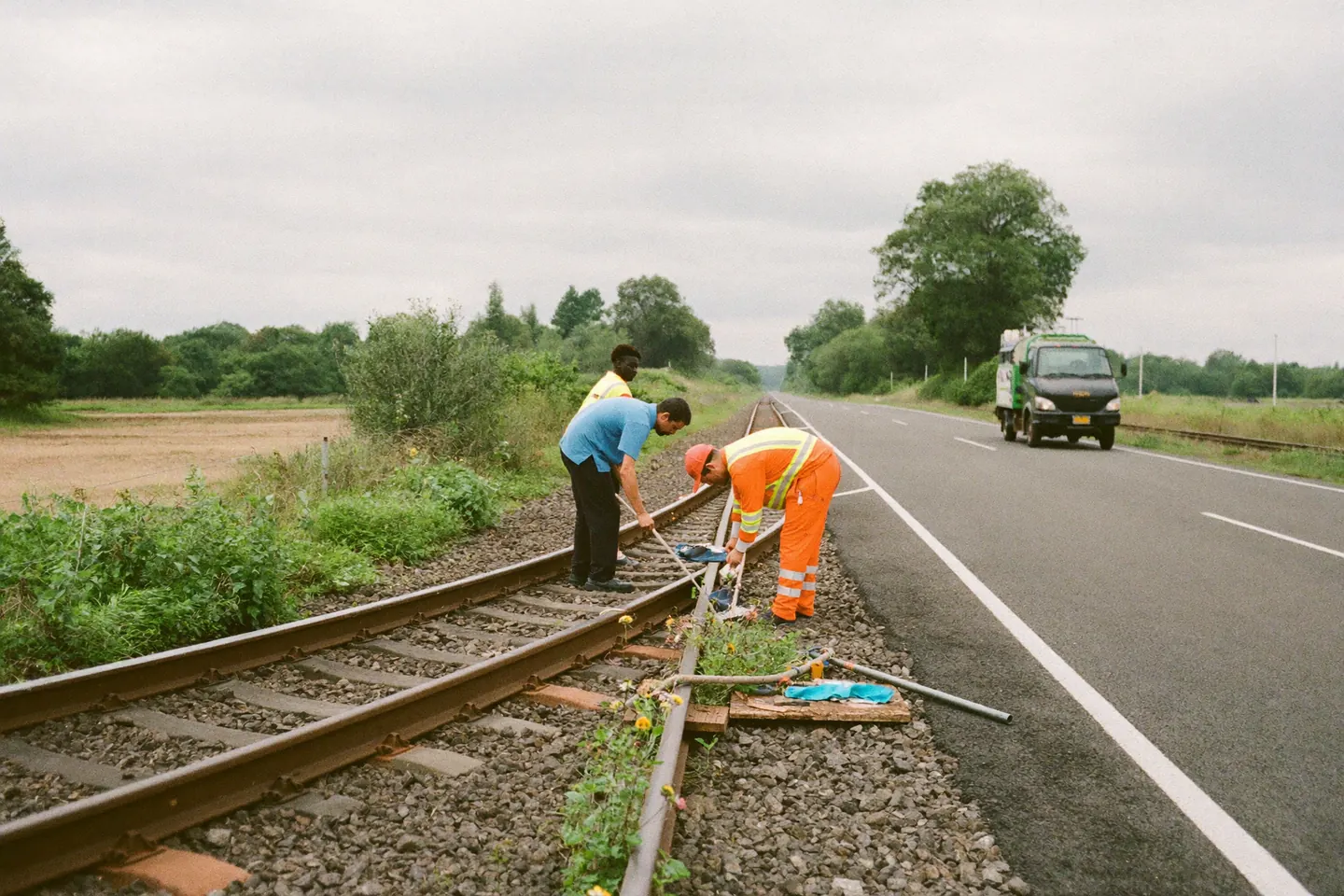 Two workers in hi-vis clothing kneel over a rail joint on a single-track rural line during a daytime maintenance window, a utility truck parked on the road alongside.